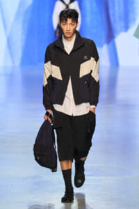 Male model walks a fashion show wearing a black-and-beige color-block windbreaker over a white striped shirt, black shorts, and carries a navy drawstring backpack.
