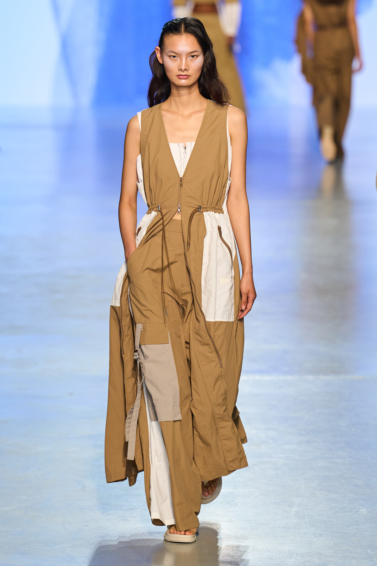 Female model walking a fashion runway in a tan and cream color-blocked sleeveless jumpsuit with a drawstring waist and pockets.