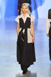 Model walking a fashion runway in a black-and-white color-block trench over a white striped shirt, hands in pockets.