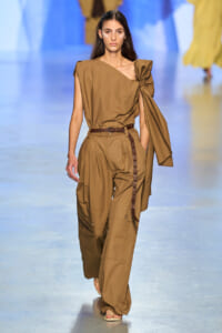 Model walks the runway in a monochrome tan outfit: one-shoulder top with a large bow and matching cargo trousers, brown belt, and sandals.