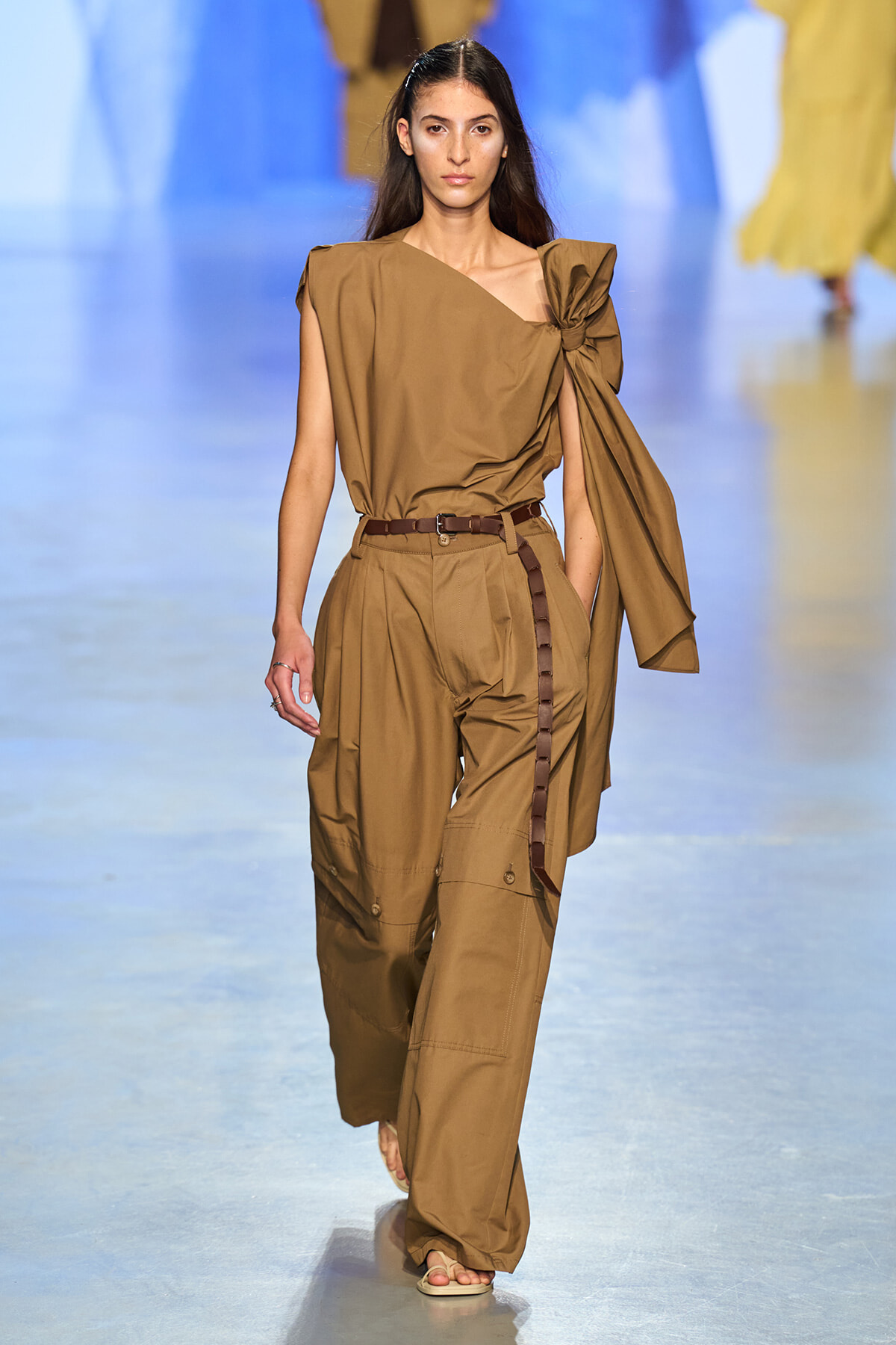 Model walks the runway in a monochrome tan outfit: one-shoulder top with a large bow and matching cargo trousers, brown belt, and sandals.