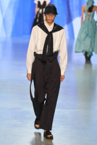 Male model on a fashion runway wearing a white shirt with a black scarf, oversized black pants, a black belt, sandals, and a black visor.