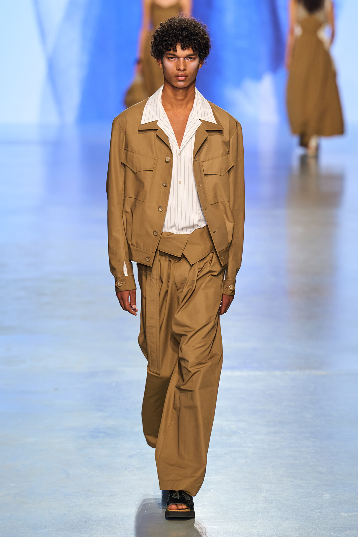 Full-body shot of male model walking runway in tan utility jacket and wide-leg matching pants over a white striped shirt at a fashion show.
