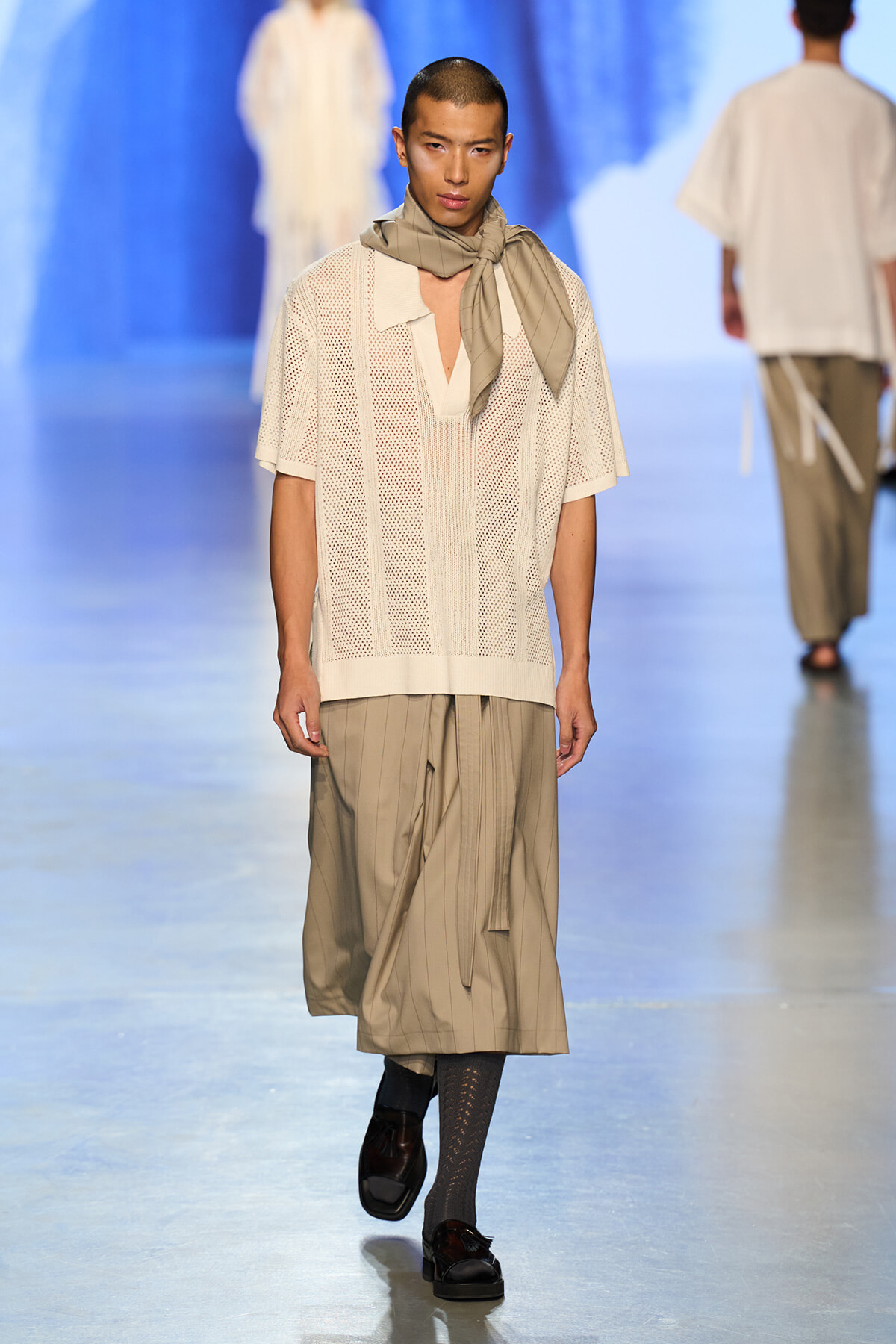Male model on a runway wearing a cream perforated short-sleeve knit top, beige pleated midi skirt, and a light scarf at the neck.