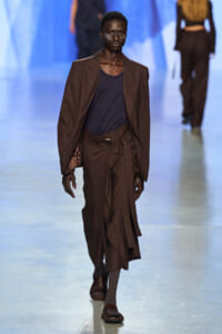 Male model walks a fashion runway in a brown pinstripe suit with a navy T-shirt, oversized blazer, and belted trousers.
