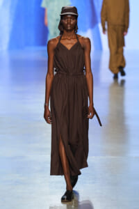 Model on a runway wearing a brown halter-neck dress with a cinched waist, matching cap, and black loafers.