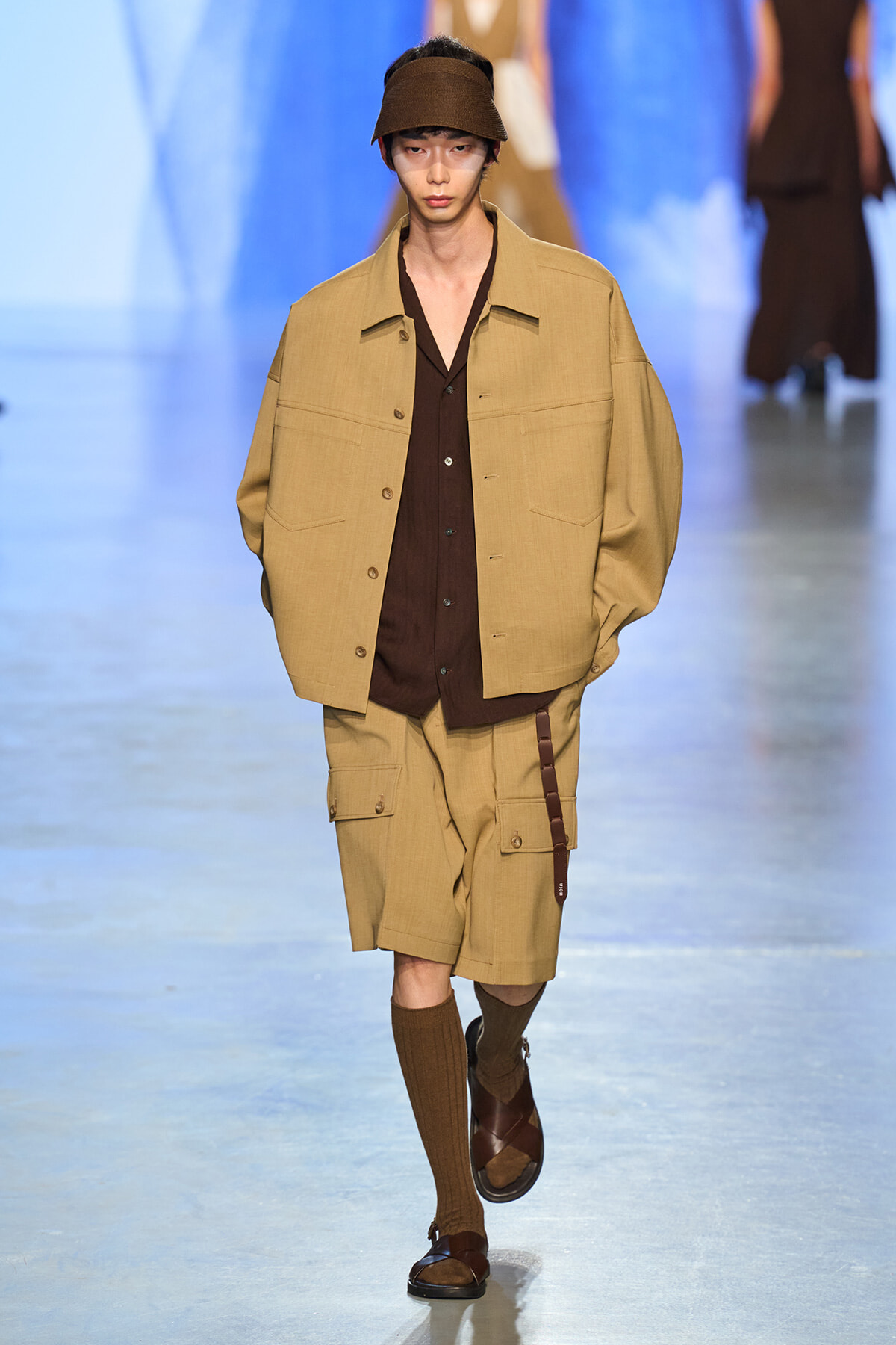 Male model walks the runway in a tan oversized jacket and matching shorts, brown shirt, knee-high brown socks, and sandals with a brown visor.