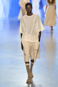 Male model in a sheer white mesh top over beige shorts with a chain, walking a fashion runway.