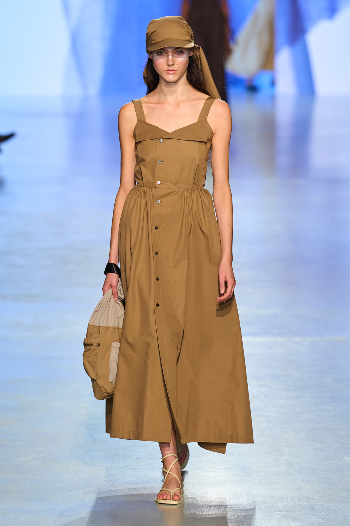 Model on a runway wearing a tan sleeveless button-front dress with a matching cap and beige tote bag in hand.
