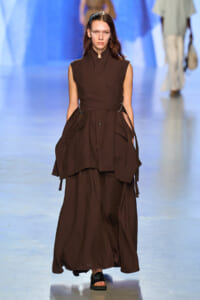 Model walks the runway in a dark brown sleeveless wrap dress with a tied belt and layered skirt.