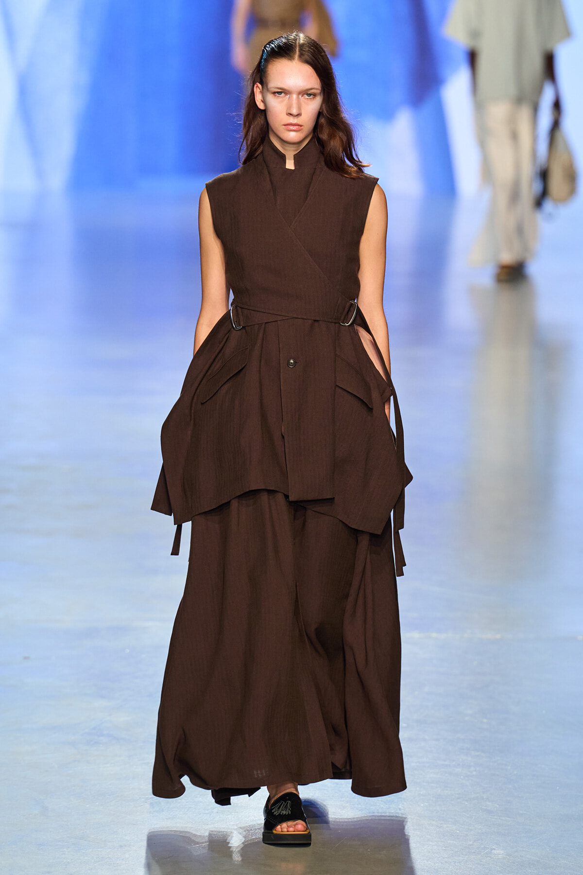 Model walks the runway in a dark brown sleeveless wrap dress with a tied belt and layered skirt.