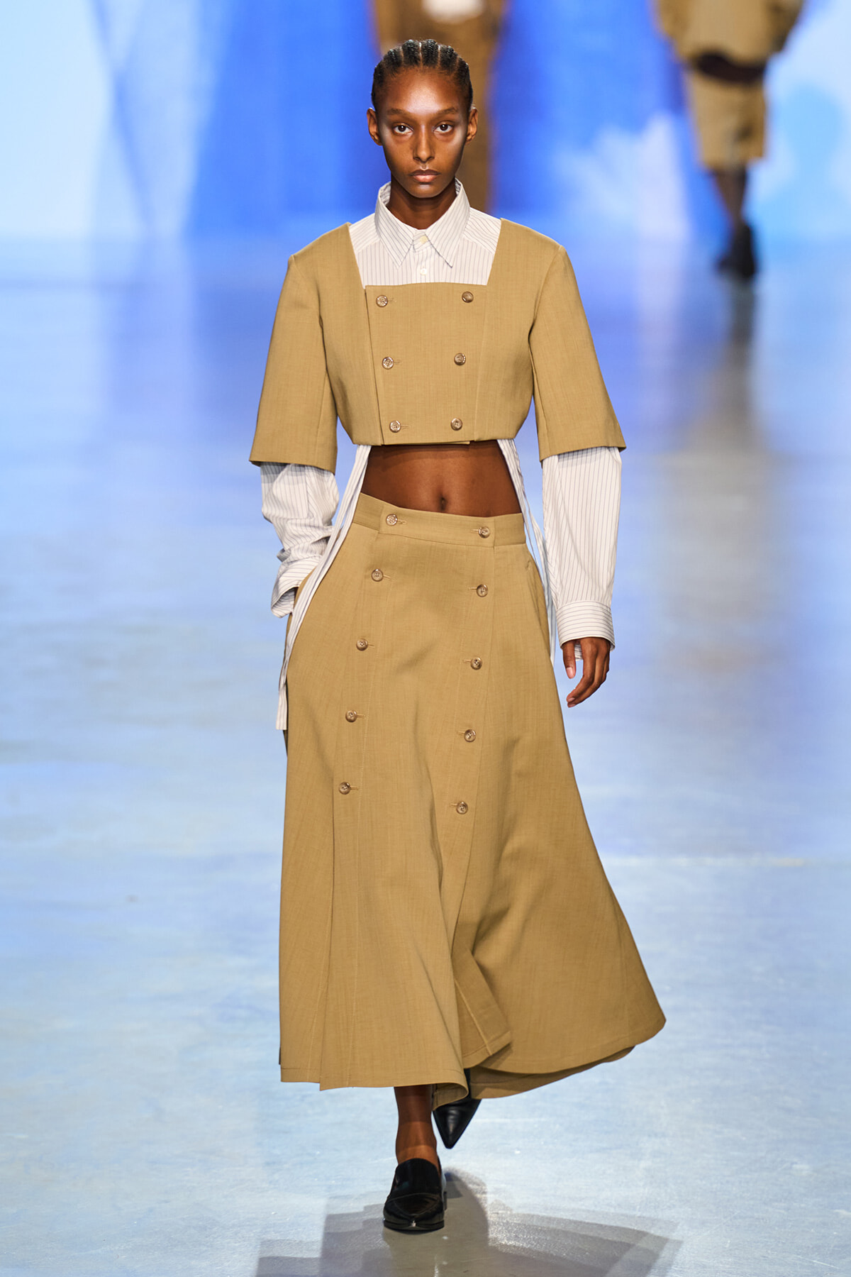 Model walks the runway in a tan cropped double-breasted jacket over a striped shirt, paired with a matching high-waisted buttoned skirt and black loafers.