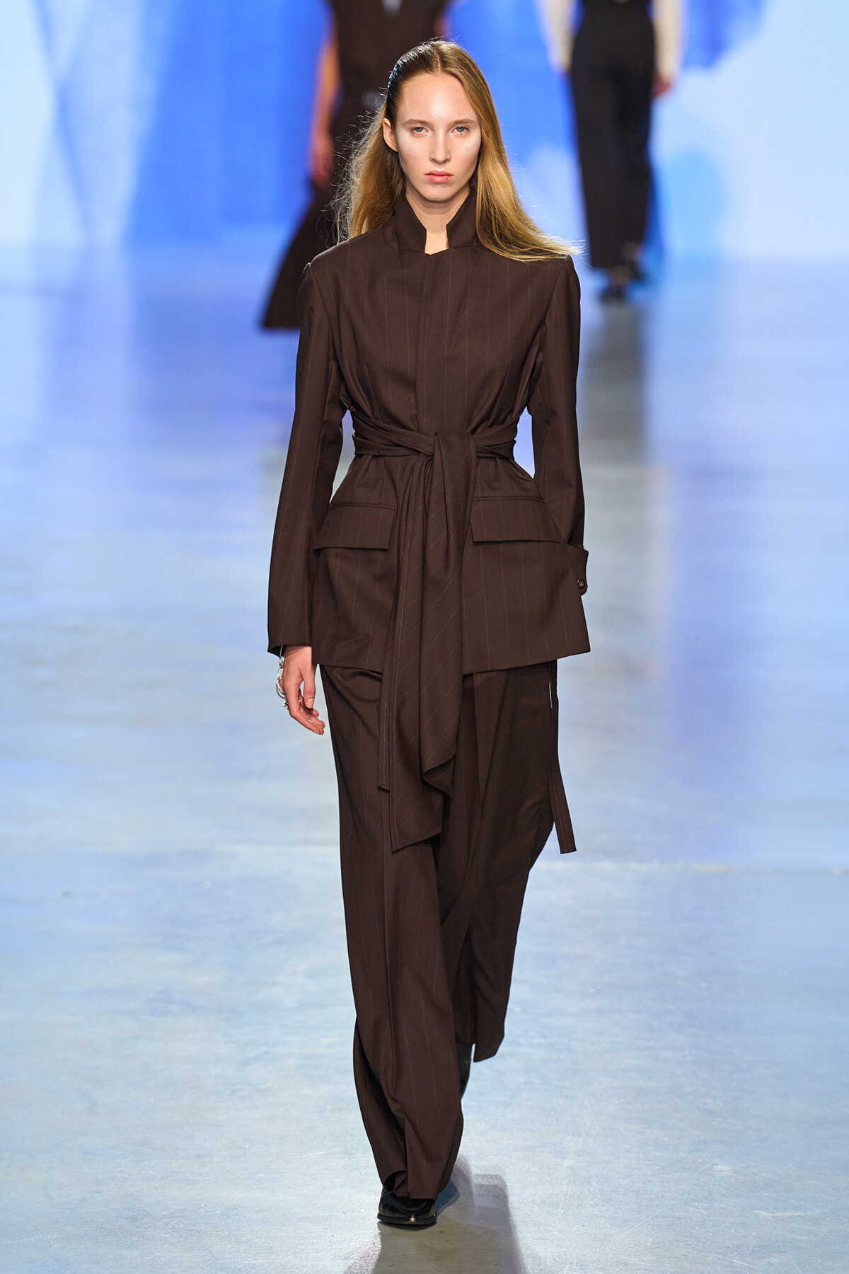 Female model walking a fashion runway in a dark brown pinstripe suit with a waist-tied belt.