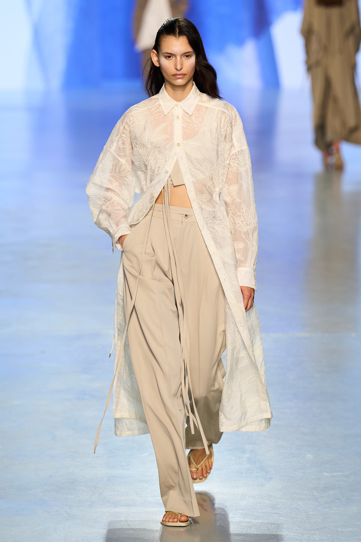 Model walking a fashion show in a sheer white lace long coat over beige wide-leg pants and sandals.