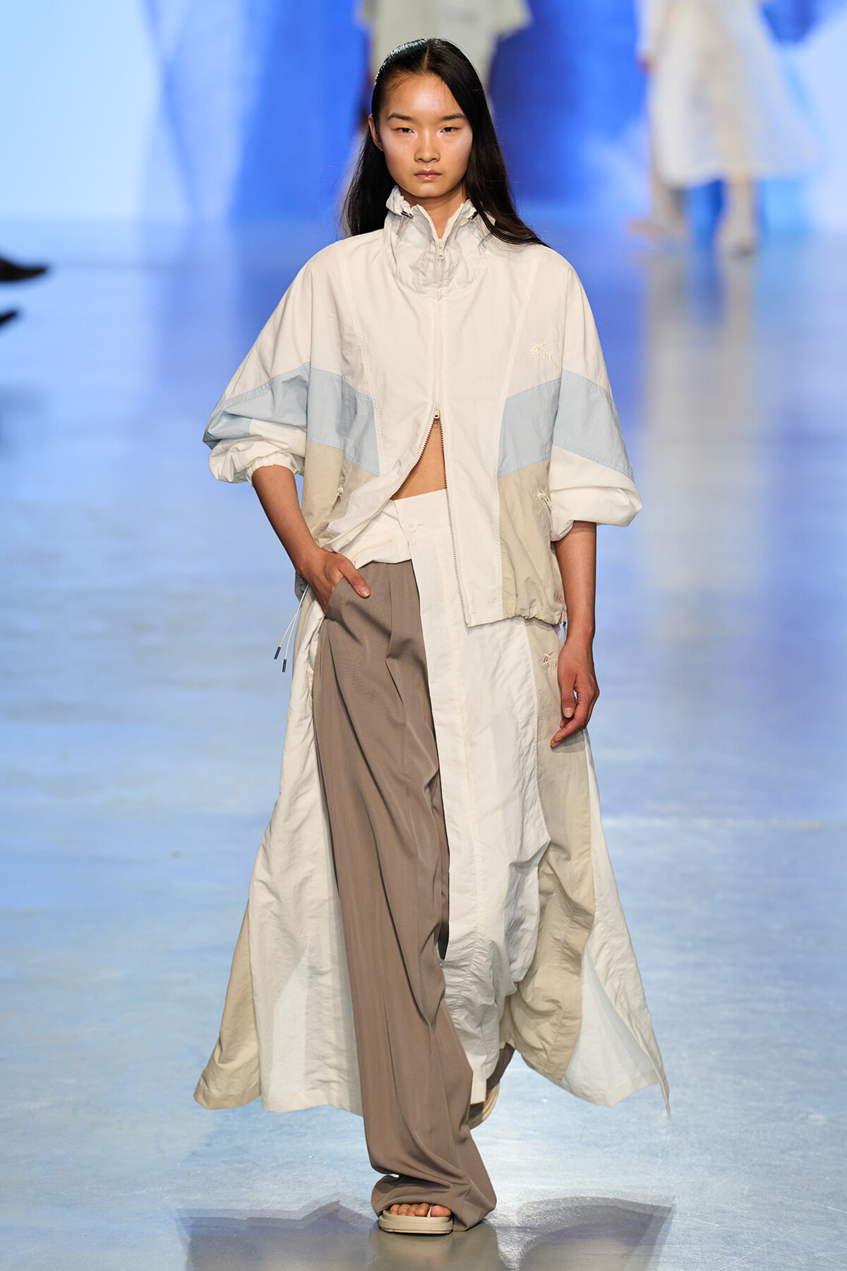 Model walks the runway in a cream color-block windbreaker with light blue panels and wide taupe trousers.