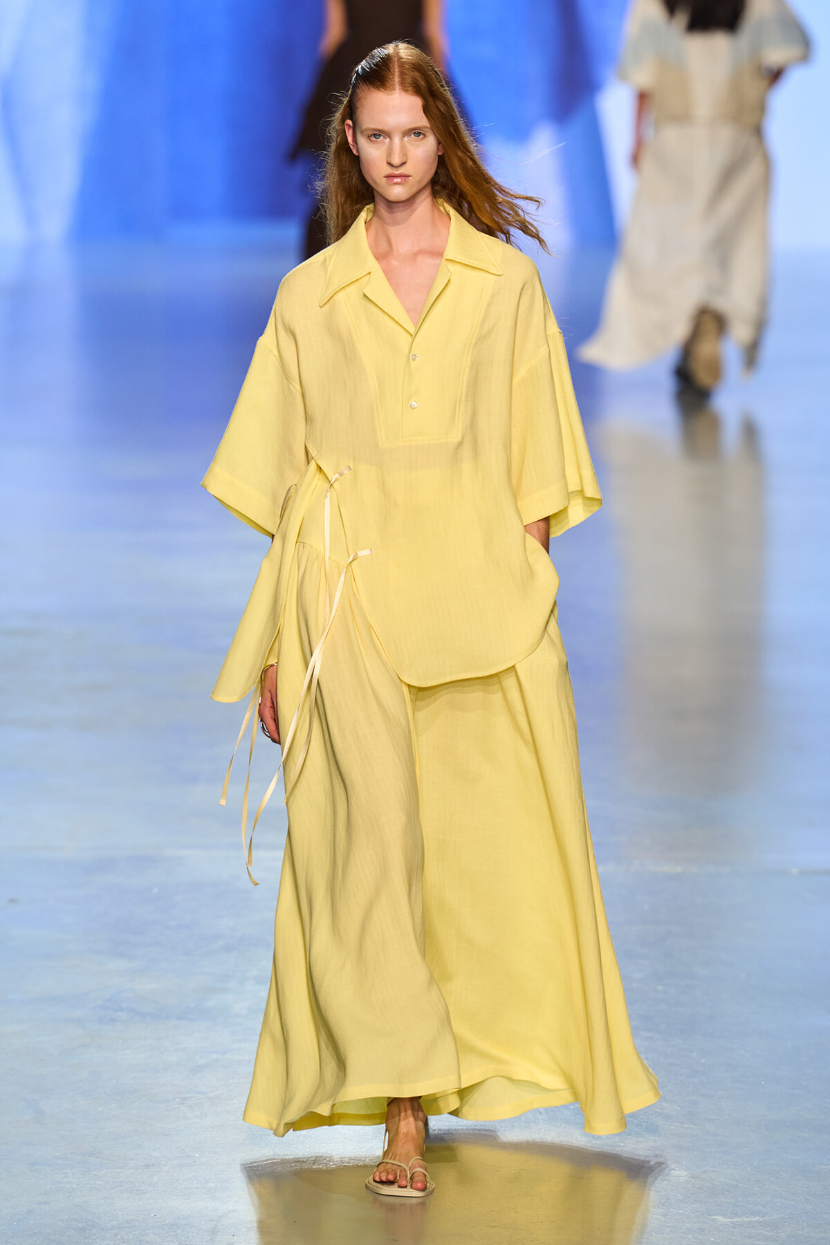 Model wearing a pale yellow loose shirt and matching wide-leg pants, walking on a runway in sandals.