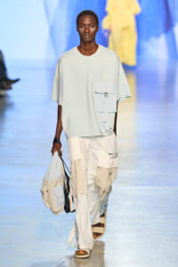 Male model on a runway wearing a pale blue oversized t-shirt with a large chest pocket, white/beige patchwork cargo pants, and a light bag in hand.