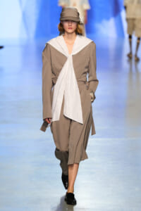 Model walking the runway in a taupe trench with a white striped scarf tied at the chest and a matching cap, black loafers visible.