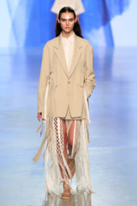 Model walking the runway in a beige oversized blazer over a white shirt with a long fringe-trimmed skirt and sandals on a fashion show catwalk.
