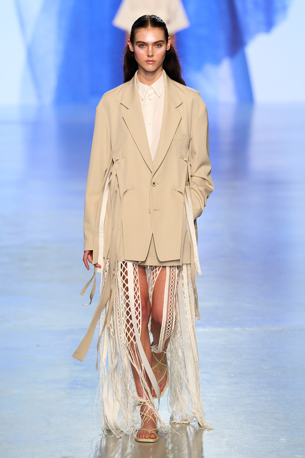 Model walking the runway in a beige oversized blazer over a white shirt with a long fringe-trimmed skirt and sandals on a fashion show catwalk.