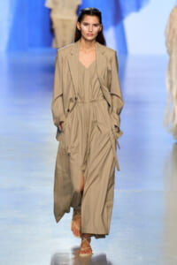 Runway model in a beige pinstripe suit with a cinched waist and open blazer, walking the catwalk in strappy sandals.