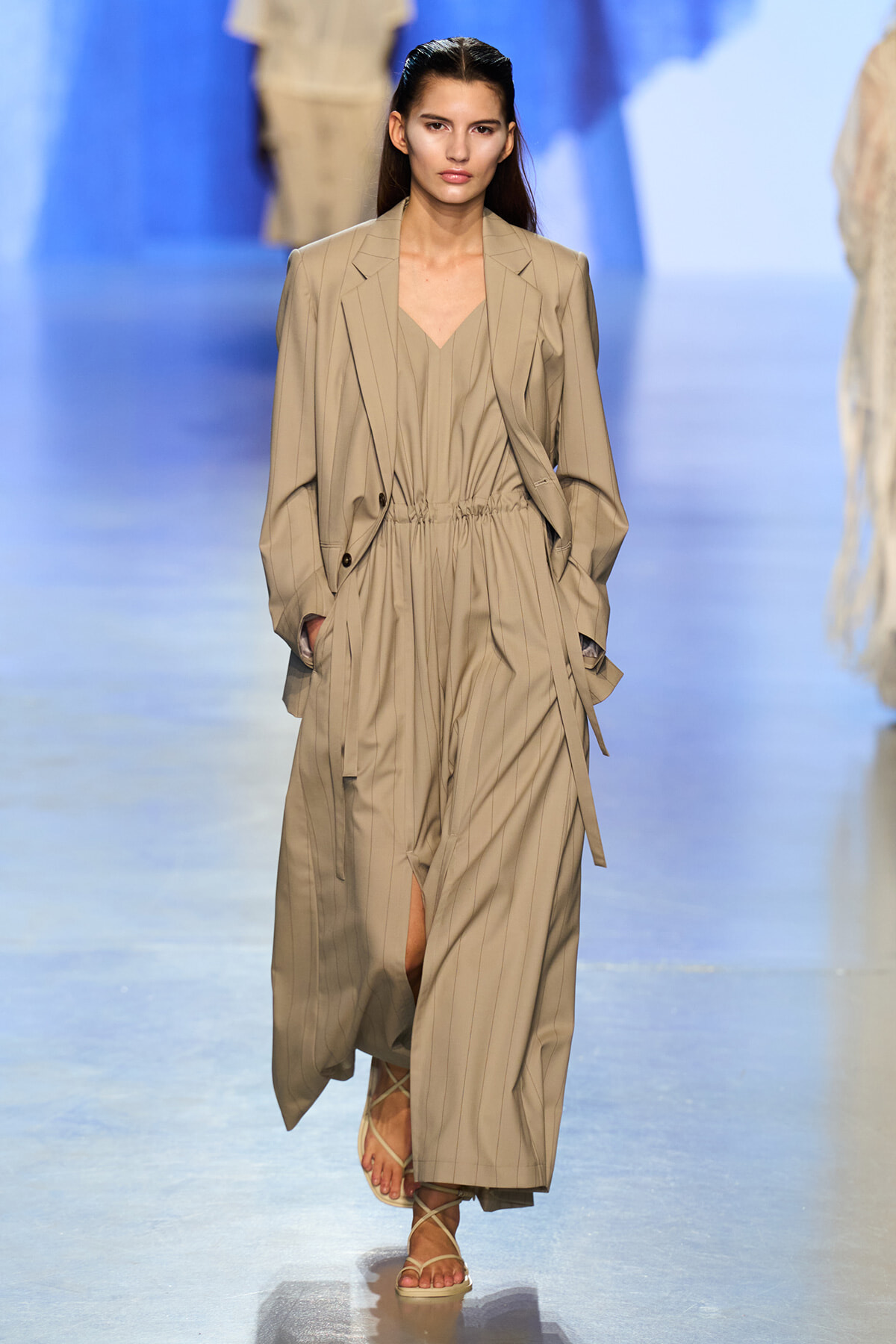 Runway model in a beige pinstripe suit with a cinched waist and open blazer, walking the catwalk in strappy sandals.