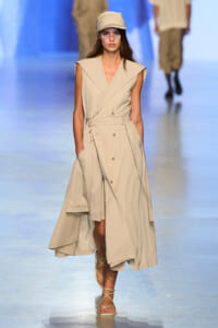 Model walks the runway in a beige sleeveless trench dress with a waist belt and matching cap; strappy sandals complete the look.
