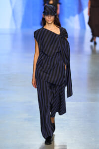 Runway model wearing a navy and black diagonal-stripe ensemble with an asymmetric top and matching wide-leg pants, plus a striped cap on a blue-lit catwalk.