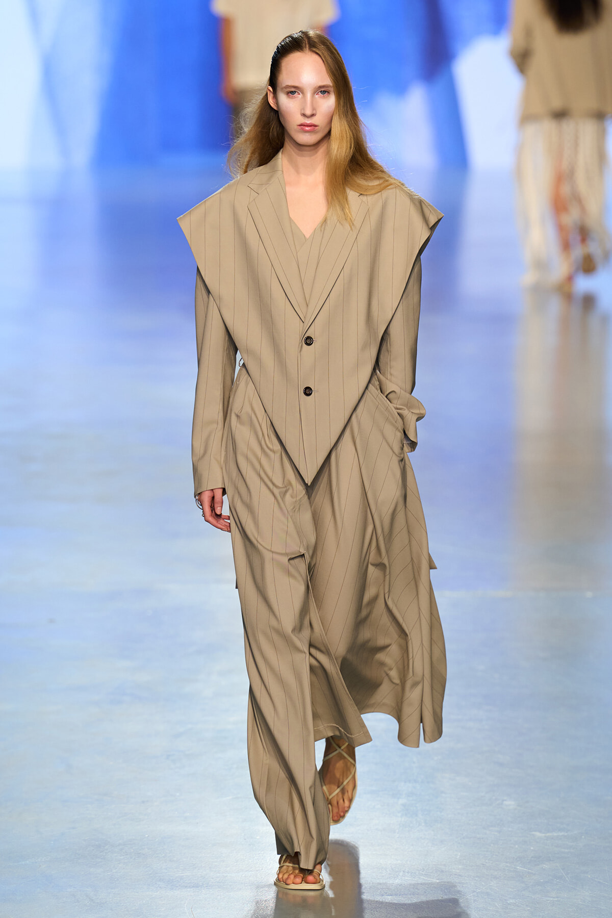 Model walks a beige pinstripe oversized suit with wide shoulders on a runway.