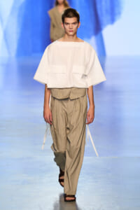 Model walks the runway in a white oversized top with large front pockets and beige pinstriped draped-waist pants, wearing brown sandals on a blue-lit runway.