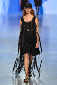 Model on a fashion runway wearing a black crochet fringe dress, black cap, and flat sandals.