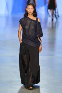 Model walks a runway in a sheer black mesh top with one-shoulder detail and wide-leg black pants with lace panels.