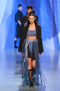Female model walks a fashion runway wearing a blue-gray crop top, black blazer, and a long fringed skirt made of net-like blue ribbons.