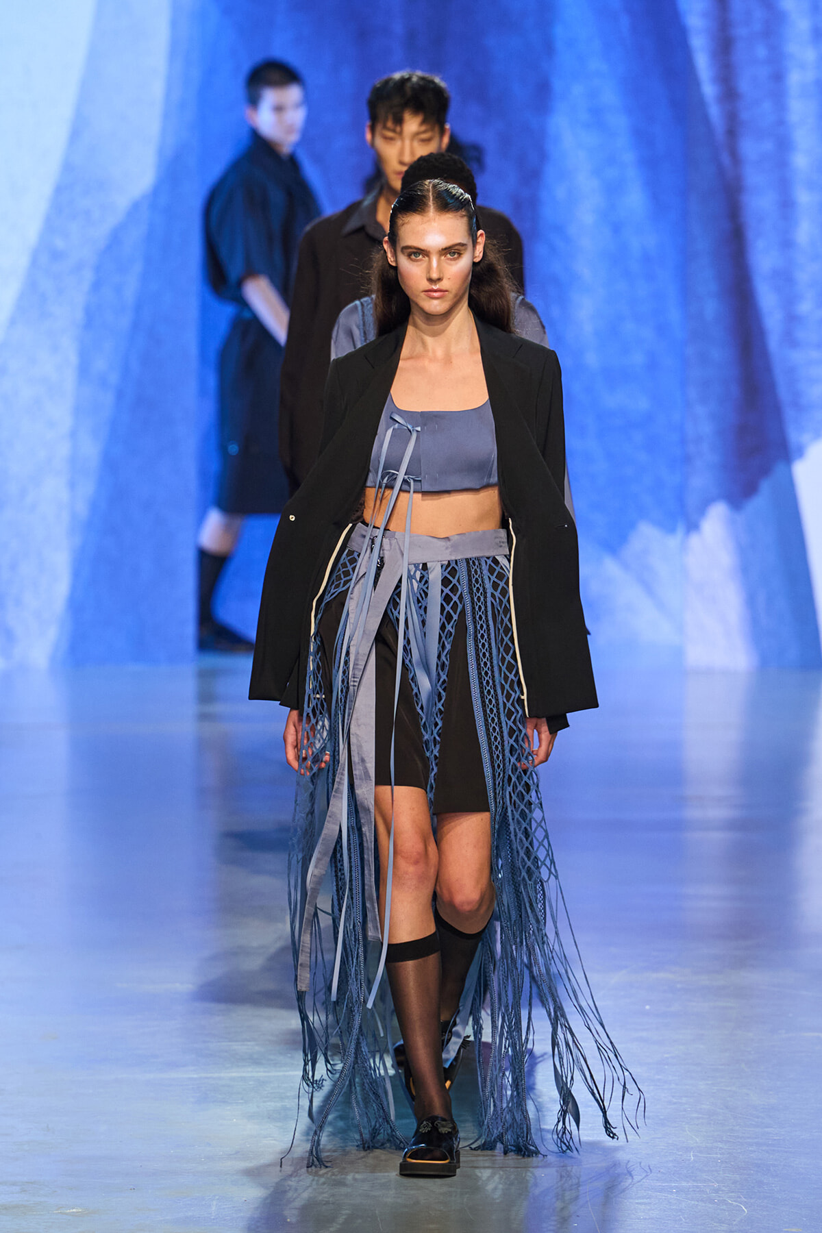 Female model walks a fashion runway wearing a blue-gray crop top, black blazer, and a long fringed skirt made of net-like blue ribbons.