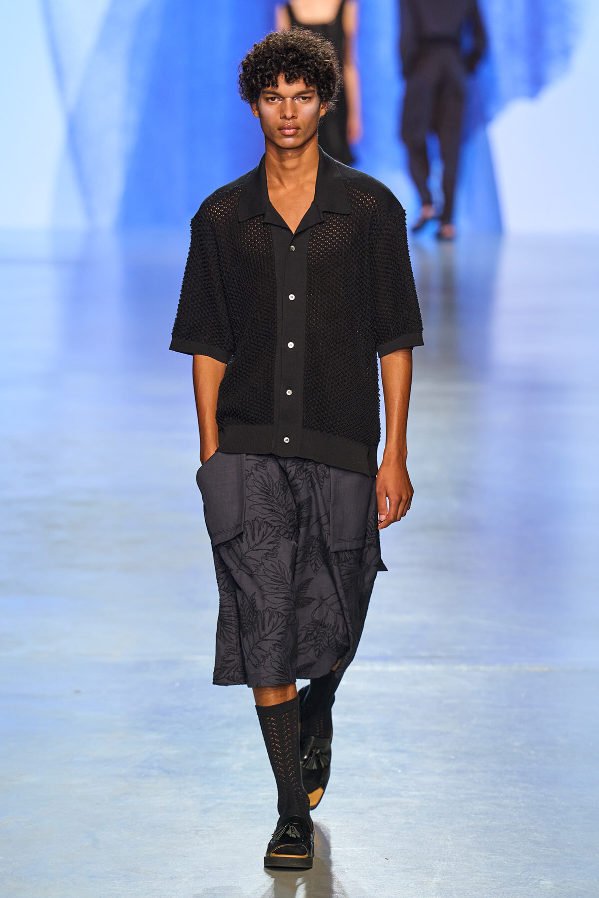 Male model walks a fashion runway in a black knit short-sleeve cardigan over a black shirt and dark patterned shorts, with black loafers and socks when he strides forward.