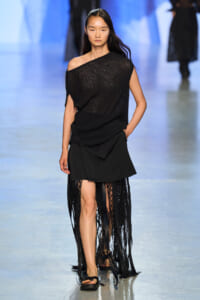 Model walking the runway in a black off-shoulder knit top and long fringed skirt with sandals.