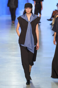 Model on a fashion runway wearing a black sleeveless top, loose black pants, a blue scarf tied at the neck, and a black cap.