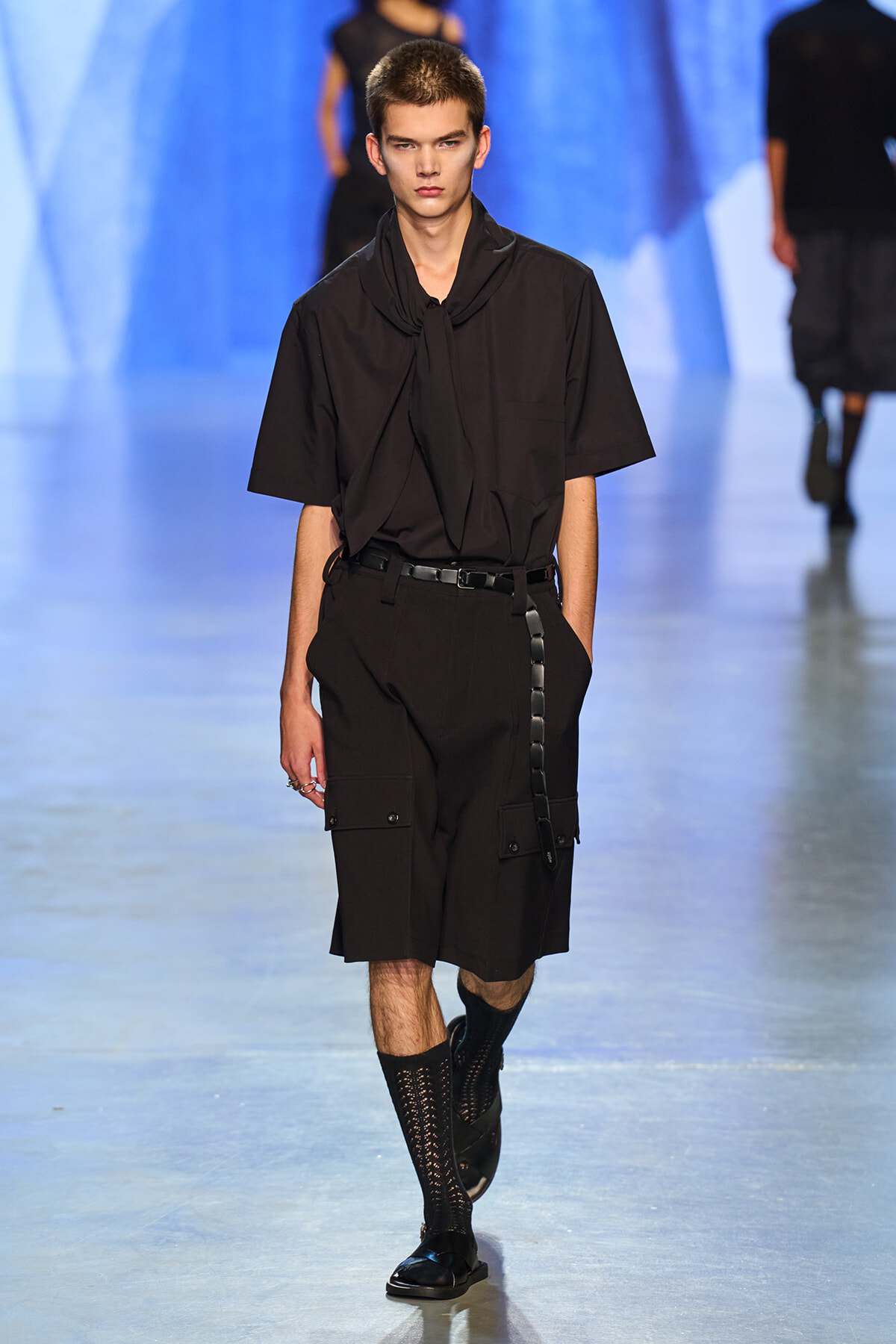 Male model walks a runway in an all-black ensemble: loose short-sleeve shirt with a scarf-like tie, cargo shorts, belt, and black sandals with socks.
