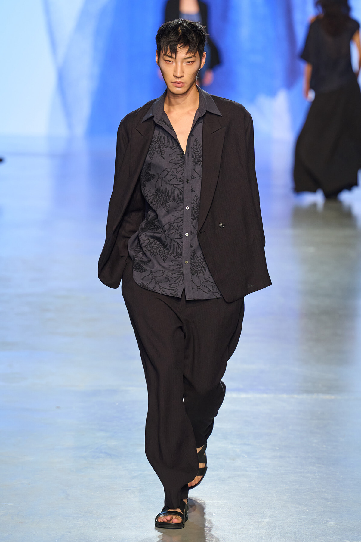 Male model walks a fashion runway wearing a dark blazer over a patterned gray shirt and loose black pants against a blue-lit backdrop.
