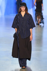 Model walking the runway in a navy and black vertical striped oversized shirt over wide-leg pants, black cap, and sandals.