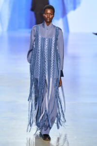 Model walks the runway in a blue-gray long-sleeve outfit with a fringed lattice overlay dress in matching tones.