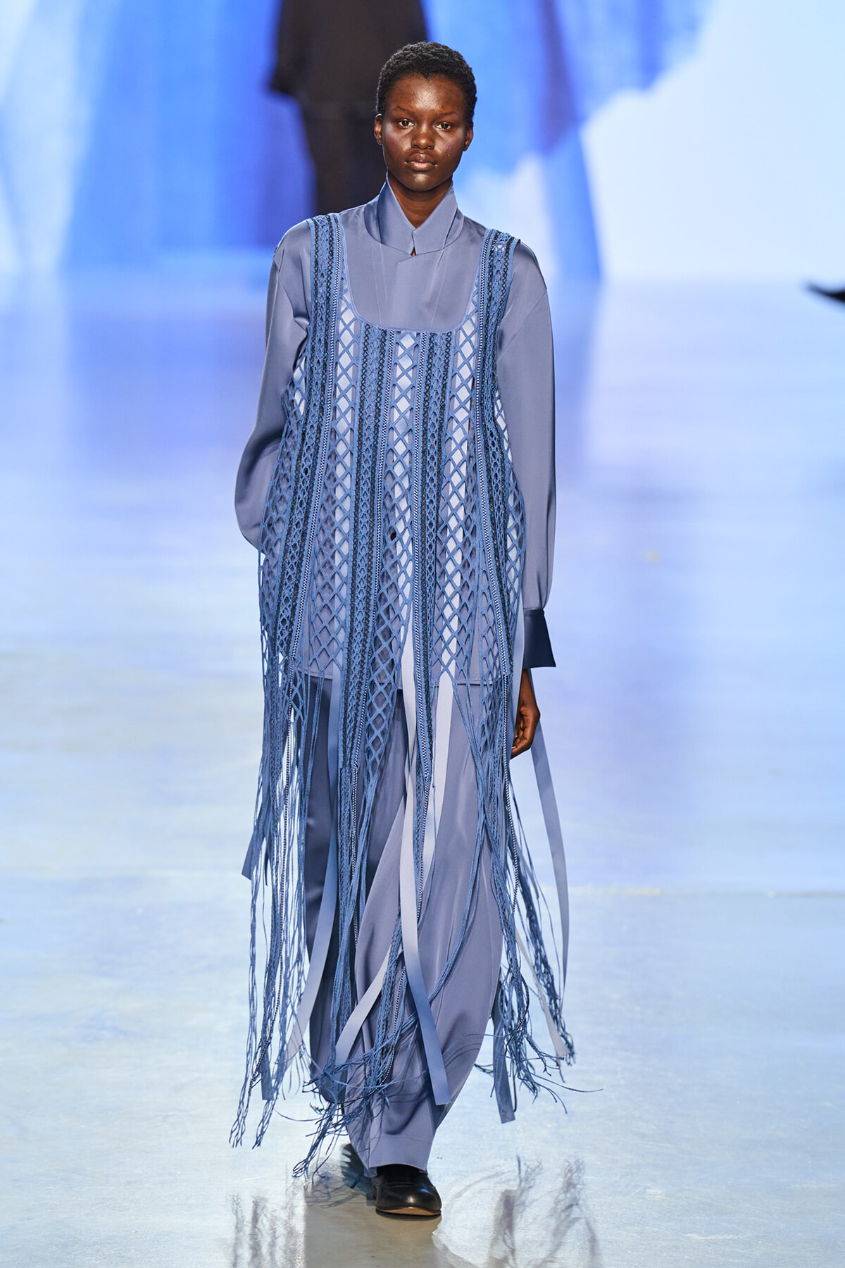 Model walks the runway in a blue-gray long-sleeve outfit with a fringed lattice overlay dress in matching tones.