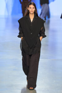 Female model walking the runway in an all-black ensemble: a voluminous blazer-like top over sheer lace sleeves, paired with loose black cargo pants and sandals, hands in pockets.