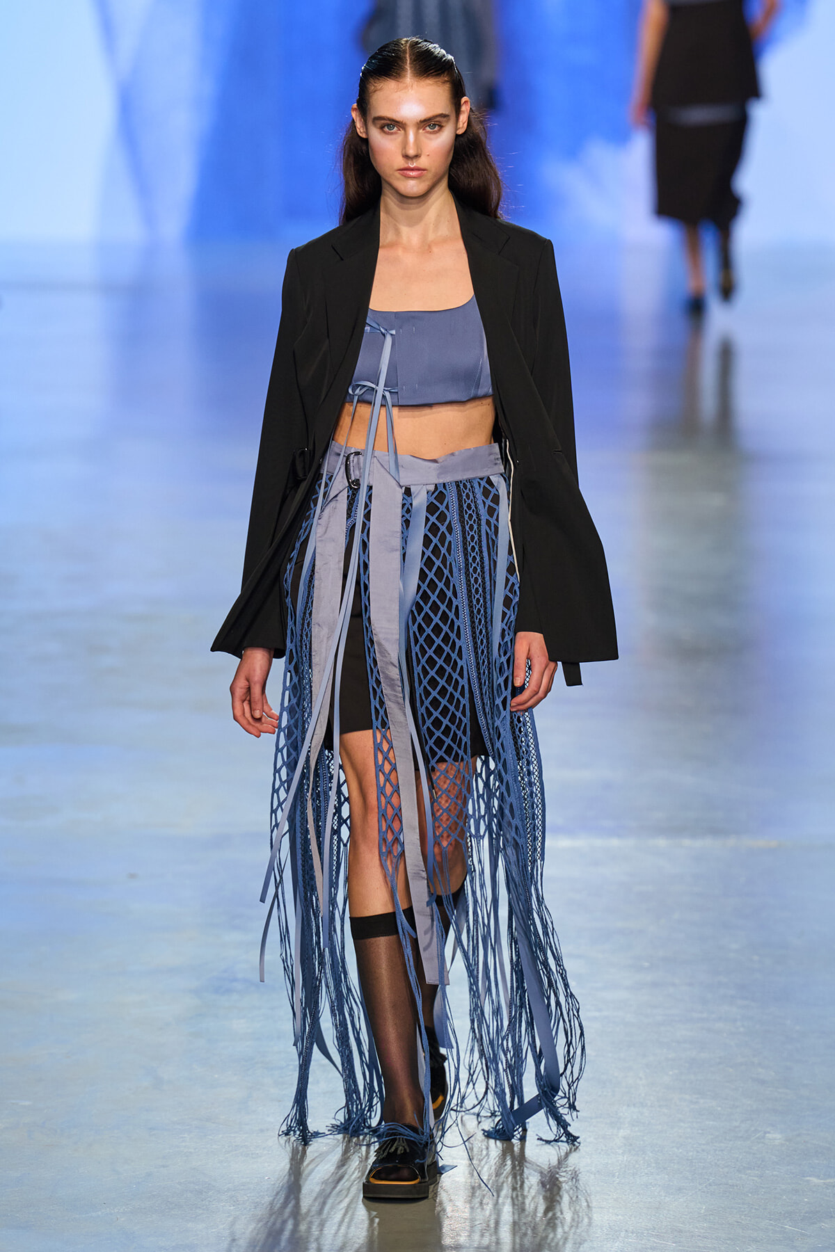 Model walking the runway in a black blazer over a blue crop top and a blue lattice-fringe skirt with long ribbons cascading to the floor.