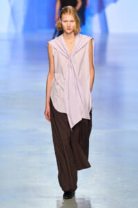 Model walks the runway wearing a lavender striped sleeveless blouse with a front tie over loose brown wide-leg pants.