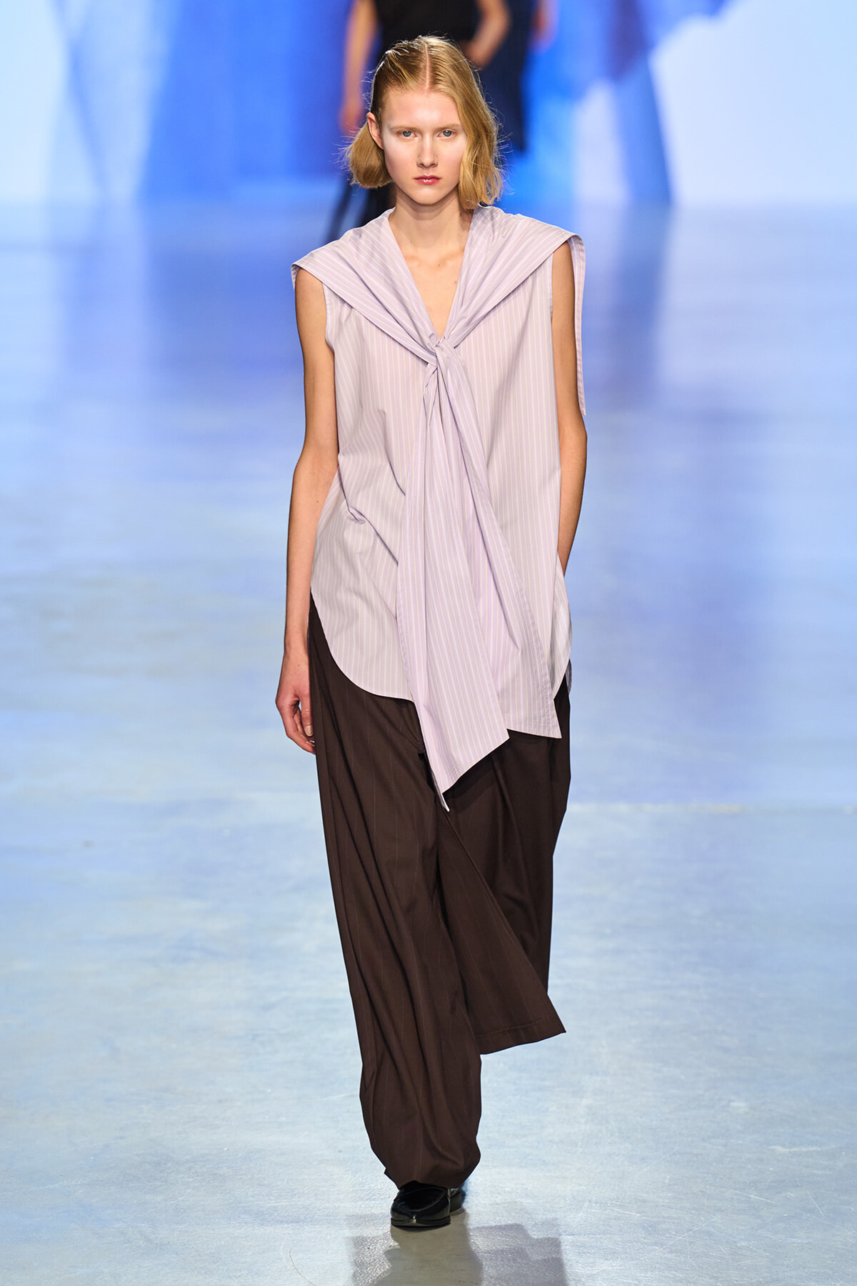 Model walks the runway wearing a lavender striped sleeveless blouse with a front tie over loose brown wide-leg pants.