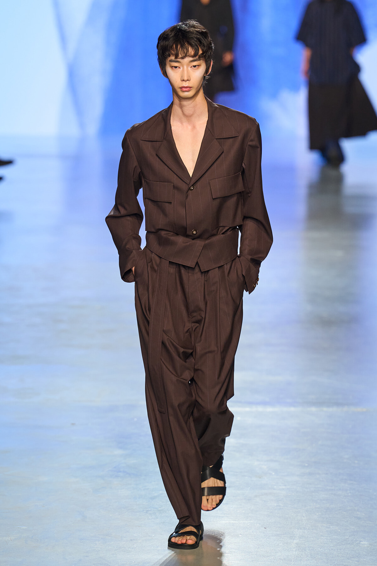 Male model walks the runway in a brown pinstriped suit with an open chest and hands in pockets, wearing black sandals.
