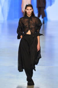 Model walking a fashion runway in a sheer black lace button-up blouse over black wide-leg pants.