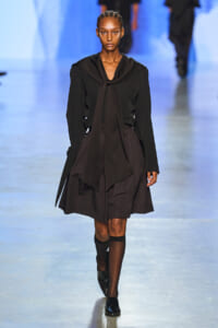 Female model walks a fashion show runway wearing an all-black layered dress with a long necktie and knee-high socks.