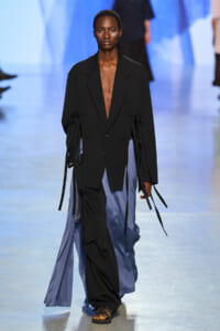 Model walking the runway in a loose black blazer with a deep V, paired with flowing blue satin pants and strappy sandals.